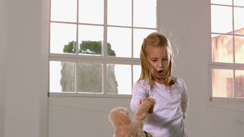 Girl in pajamas jumping and swinging teddy bear in front of window in slow motion