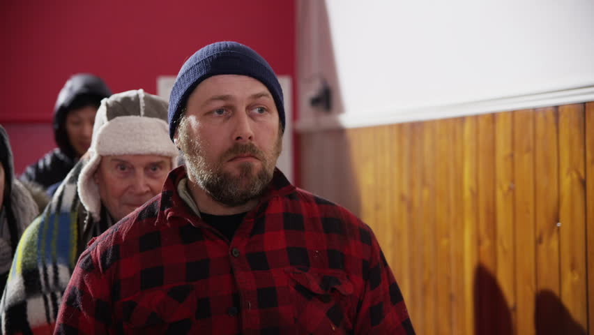 A group of homeless and needy people, wrapped up warm against the cold outside are standing in line in a refuge or soup kitchen. In slow motion.