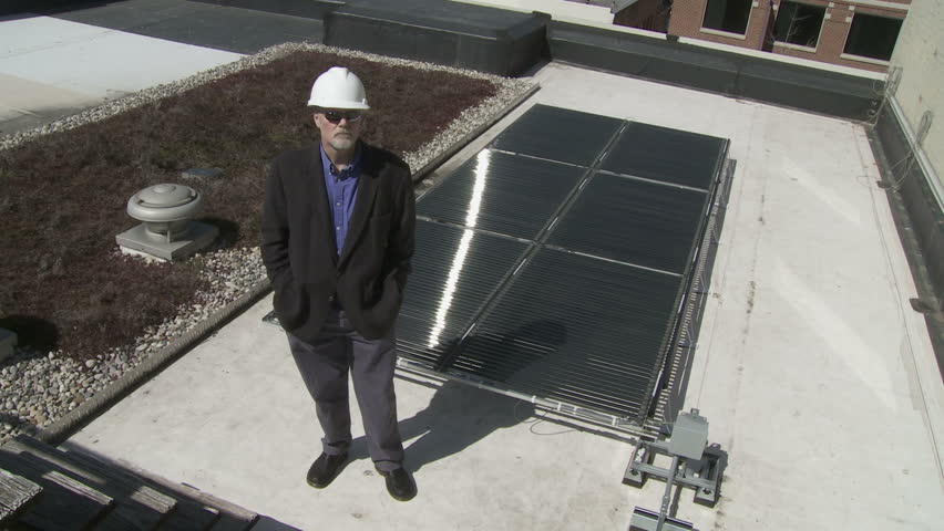 Technician stands in front of a solar power installation on a roof top. This could be a building contractor, workman, architect or site inspector, wearing sunglasses and a hard hat. Wide shot.