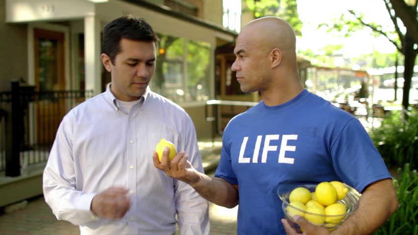 Man in a blue T-shirt that says "LIFE" hands lemons to people on the street. Man takes one and it changes (jump cut) to a jug of lemonade.