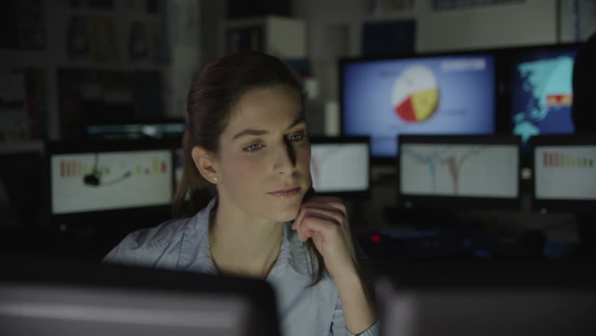 A young business woman is working late at night in a dark office filled with computer screens. She is feeling tired and overworked.