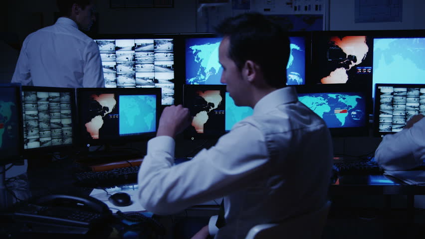 A team of security personnel man the stations within a busy system control room. Could be a weather or power station or air traffic control. It could be a  police/army control facility. 