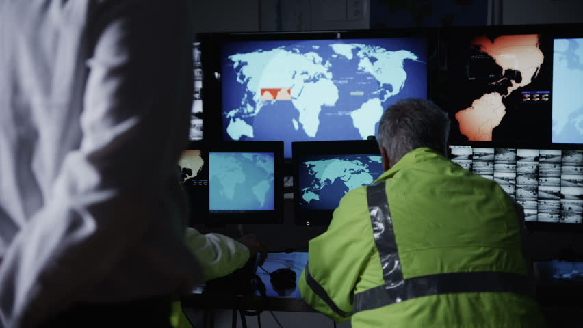 A team of security personnel working in a busy system control room, could be a weather station/airport traffic control. It could be a  power station or police/army control facility. In slow motion.