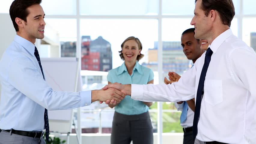 Businessmen shake hands while other business people applauding in a bright office