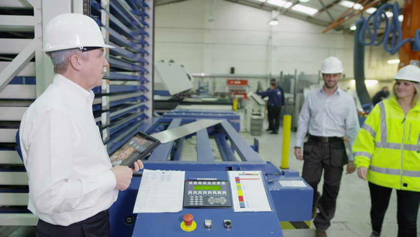 Male and female workers within a factory or warehouse are being shown by their foreman how to operate some heavy machinery.