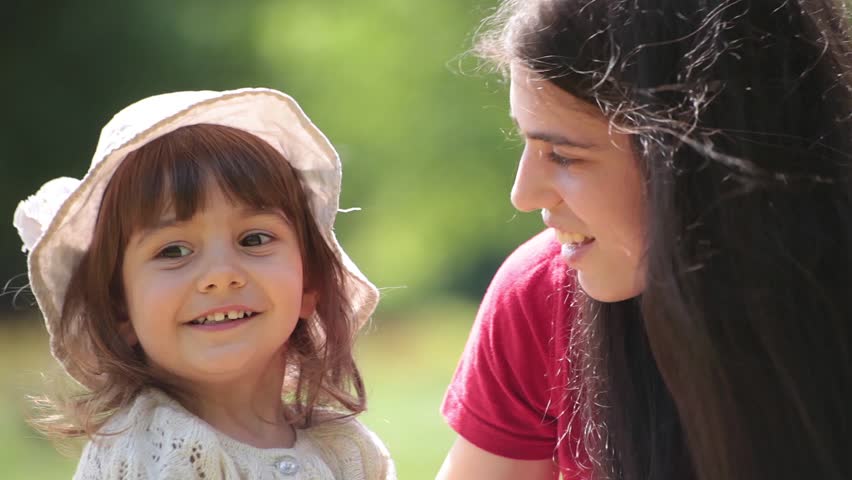 Brunette young adult mother talks to her little baby girl. Sunny day, outdoor portrait.
