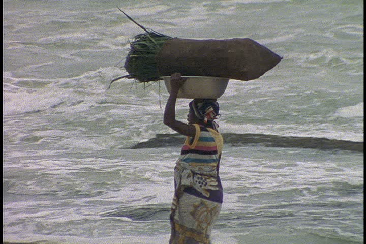 GHANA, AFRICA - OCTOBER 24, 1996: MS woman walks along the beach carrying a bundle on her head. Waves and surf crash on shore.