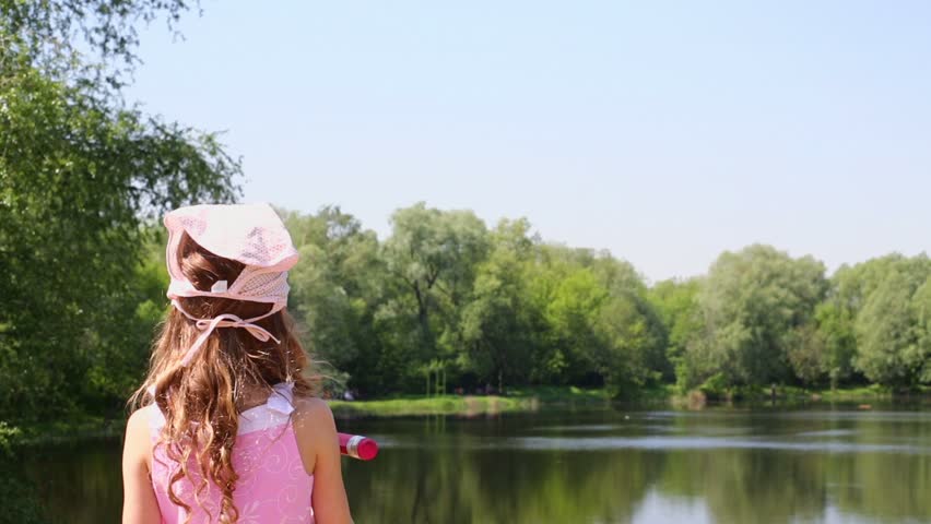 Girl imitates writing with large red pencil near pond in park at sunny summer day