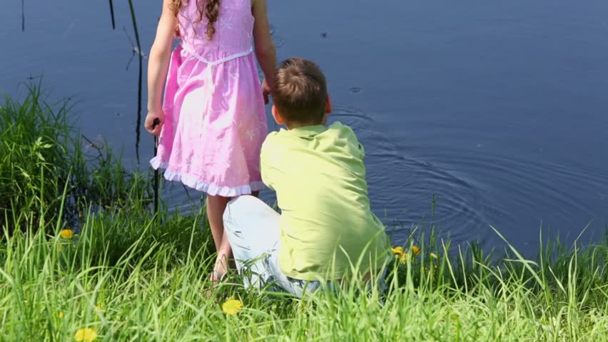 Two kids boy and little girl sit near pond and play with fishing rods