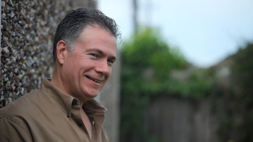A man leaning up against a tall stone fence, grins, chuckles, and the gazes contentedly at the camera.