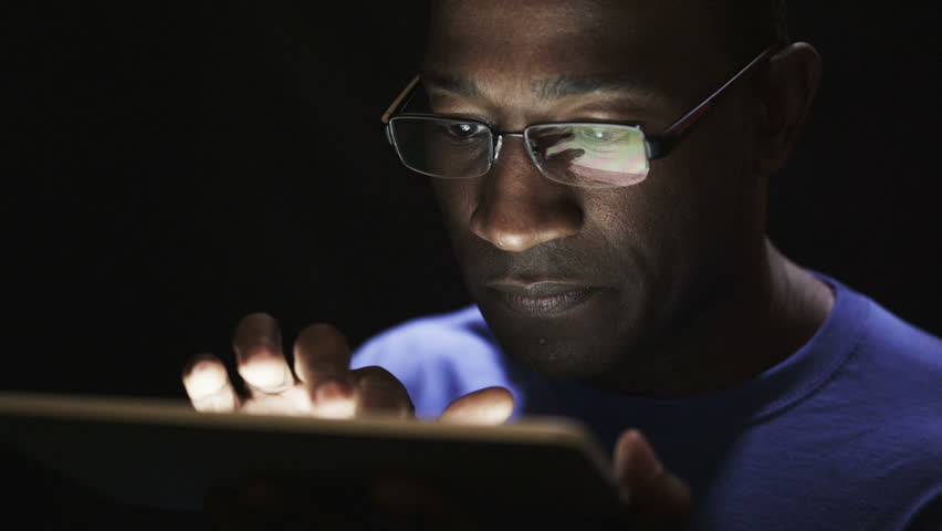 Close up of young black man reading his tablet in the dark. Black background. 