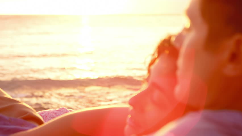 A woman lays on a man's chest as they sit on the beach and watch the sun set together