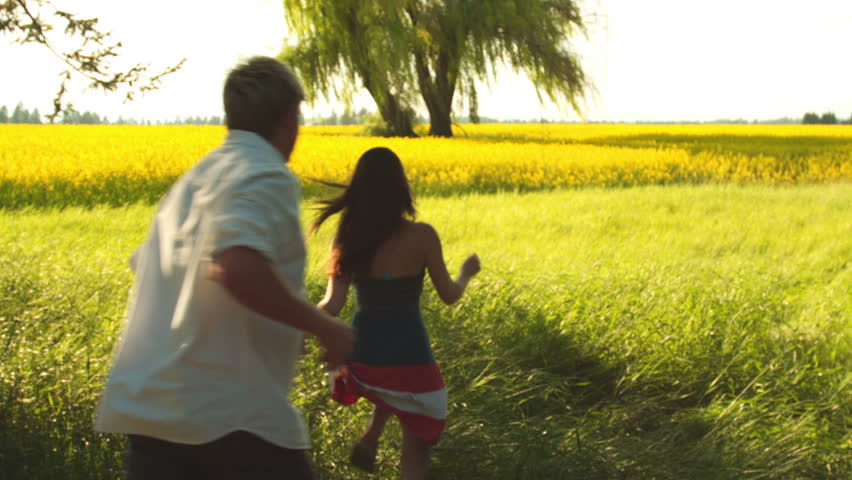 A young couple holds hands as they run through a wide open yellow field
