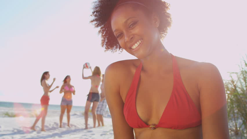 Close up pretty young African American girl smiling to camera sitting beach friends playing ball background sun lens flare shot on RED EPIC, 4K, UHD, Ultra HD resolution