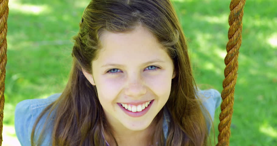 Smiling little girl sitting on a swing in the park looking at camera on a sunny day