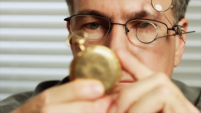 A man wearing jewelers glasses, picks up an old pocket watch and begins to examine it.  Rack focus