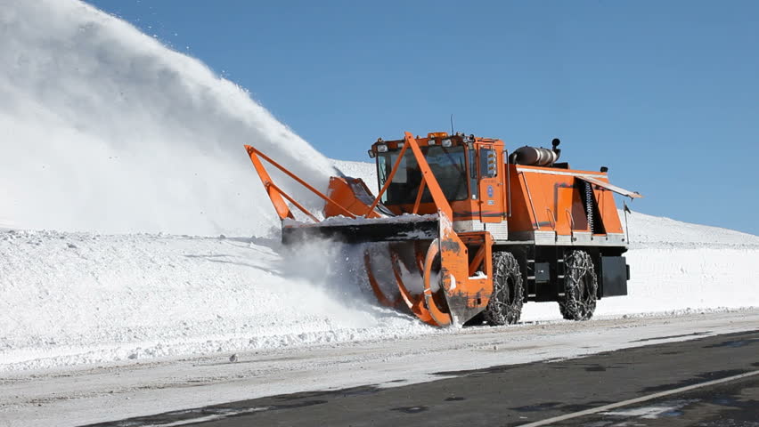 Snow blower on high elevation mountain road removing snow. Large vehicle with spinning blades throws ice and snow off the road. Deep and dangerous snow.