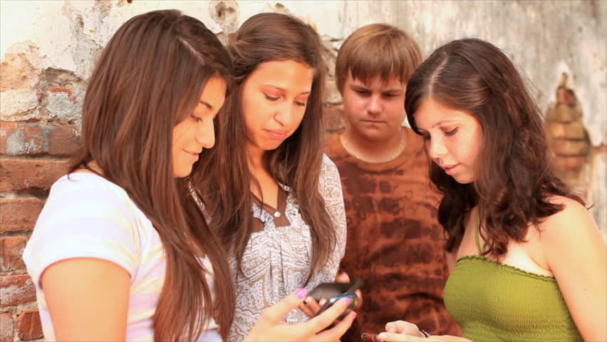 A group of teenagers hanging out by an old brick wall, using cell phones, and talking.