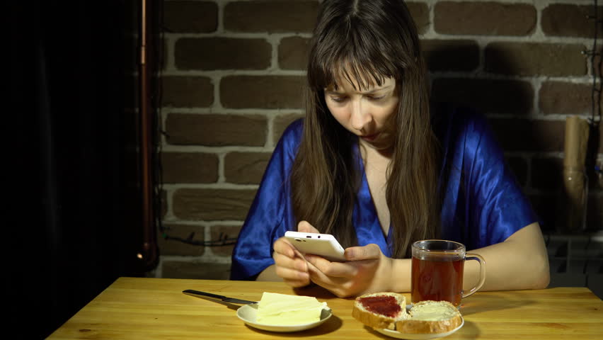 A Young Sleepy Woman In A Blue Robe Browses The Video On Her Smartphone And Drinks Tea Sitting Next To A Brick Wall 4k A Young Sleepy Woman In A Blue Robe Browses The Video On Her Smartphone And Drinks Tea Sitting Next To A Brick Wall 4k