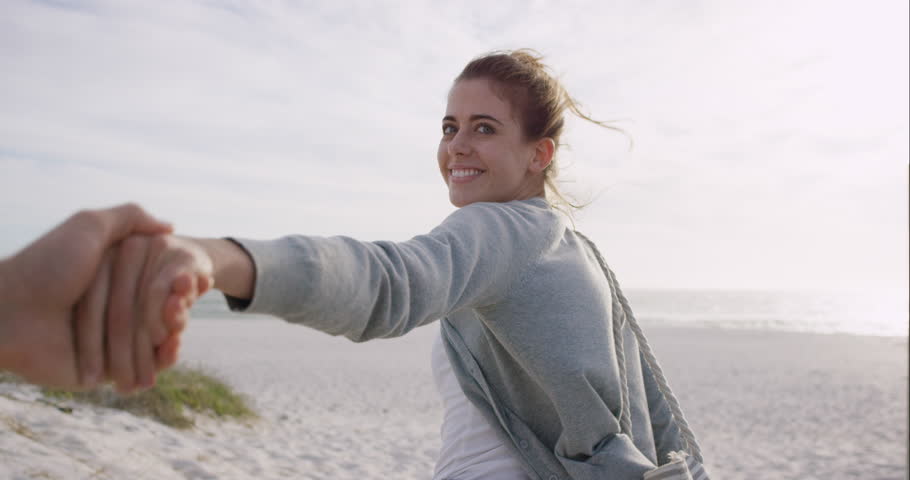 Young couple holding hands woman leading boyfriends walking towards sunset on empty beach POV travel concept