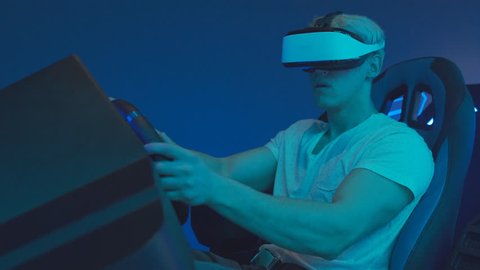 Young Man Gamer Wearing Virtual Reality Headset Plays Online Video Game With Racing Simulator