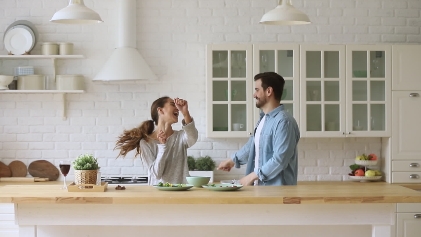 Young happy active family couple dancing laughing together preparing food at home, carefree joyful husband and wife having fun cooking healthy romantic dinner meal listen to music in modern kitchen