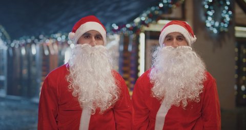 4k Portrait Of Two Santa Clause After Work Or Giving Gifts Two Happy Santa Claus Twin Brothers Removing Hat Fake Beard Near The Wooden Cottage Funny Twin Santa Clauses Giving A High Five