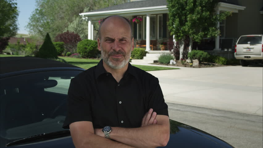 A Grey Bearded Man Leans Against Car In Tidy Suburban A Grey Bearded Man Leans Against Car In Tidy Suburban Neighborhood