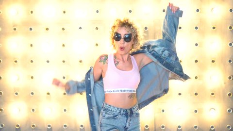 Young sexy woman in denim with short curly blonde hair dancing against a wall of light bulbs.