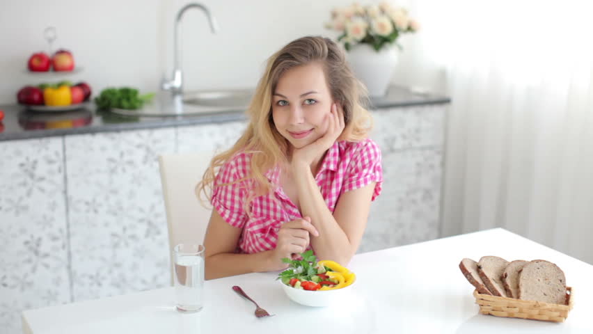 Eat glass. Cute girl sitting at Kitchen Table eating Fruit Salad and looking at Camera with smile. Woman with perfect healthy Fresh Skin sits at the Table.